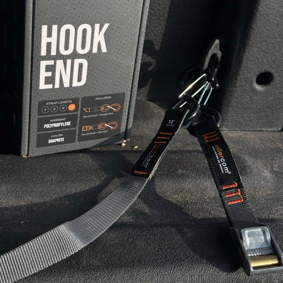 A Rolleraam Hook End strap shown with its black retail box and the grey polypropylene strap attached to a metal D-ring in a dark truck bed.