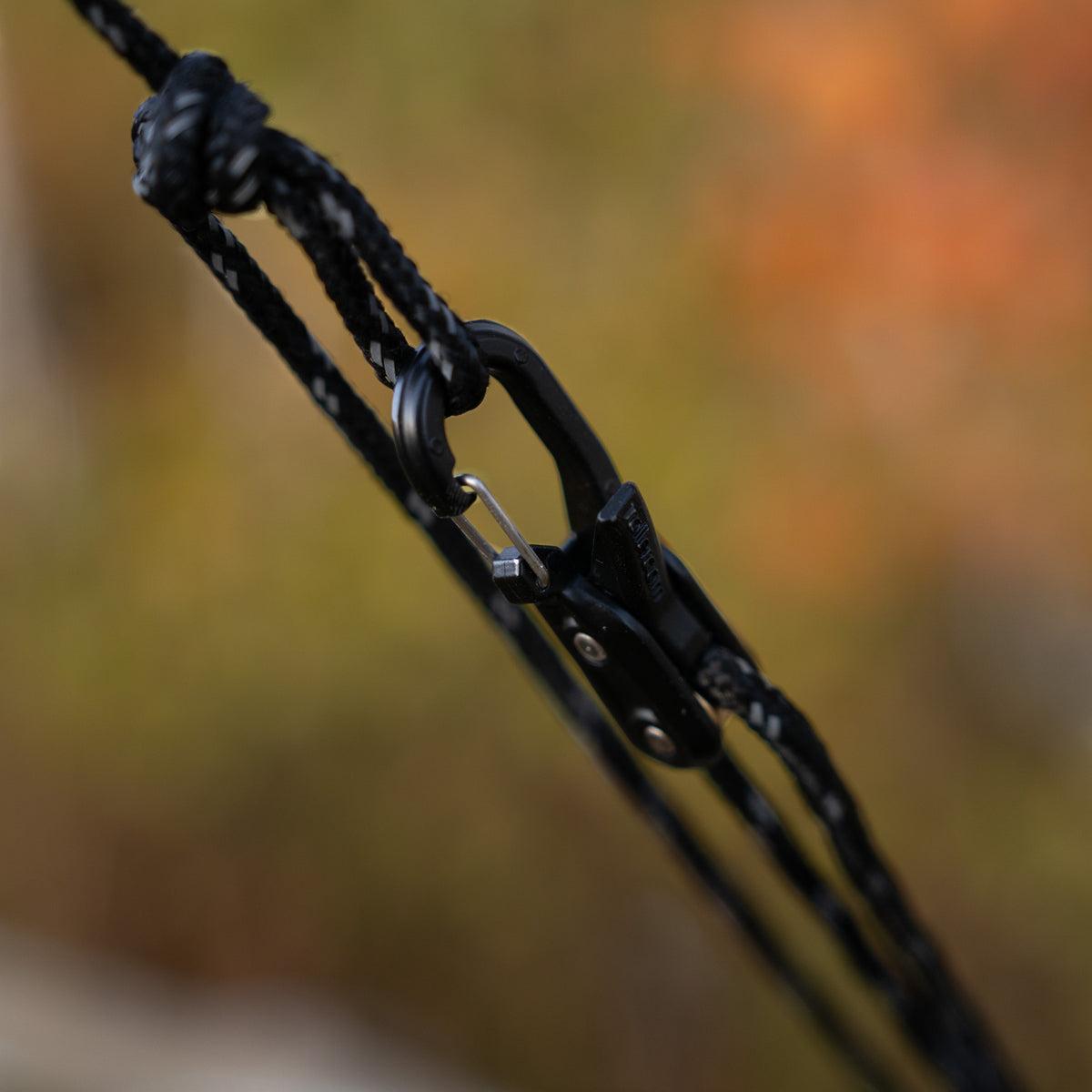 Close-up of a black Stealth Roperoller clipped onto black accessory cord, with a blurred outdoor background in warm autumn tones.