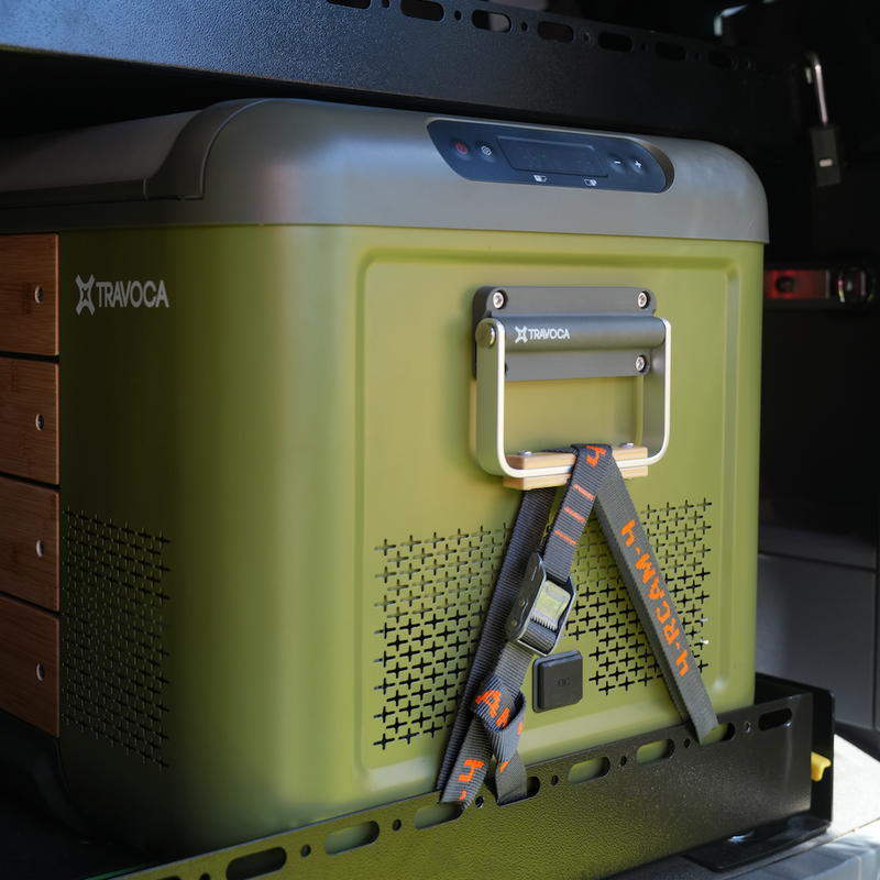 A close-up of a green electric fridge, mounted inside a vehicle compartment. The cooler features a carrying handle, ventilation grilles, and a black and orange Rollercam 4ft strap secured to the front.