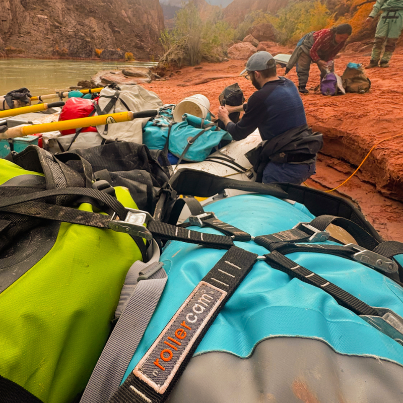 Rollercam Spider System securing pile of dry bags on raft, with a group of people preparing for a river rafting trip in a canyon with kayaks and backpacks in the background.