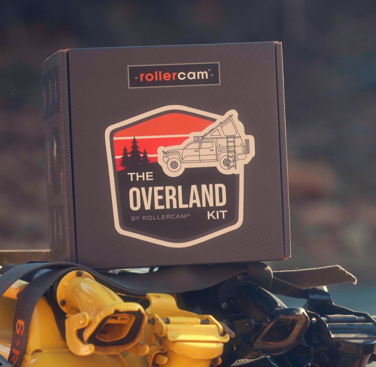 A dark charcoal gray Rollercam 'Overland Kit' box is shown resting on yellow and black gasoline jerry cans in an outdoor, blurred natural setting. The box features a prominent graphic of a vintage truck against a red and orange sunset with pine trees.