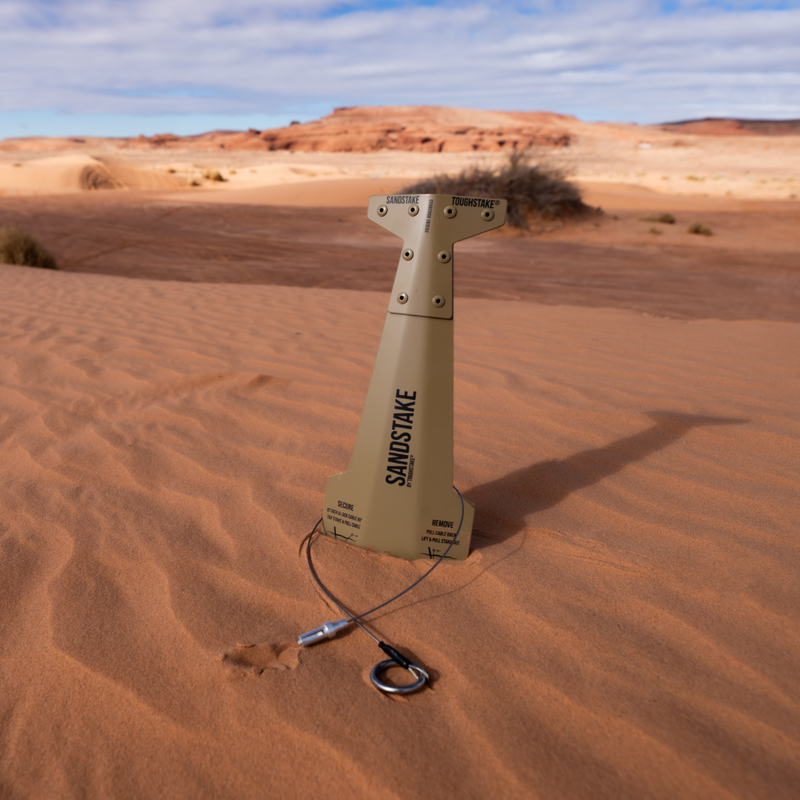 Sandstake standing upright in a desert sand dune, with a stainless steel guywire attached at the base. The background shows a blurred desert landscape with red rock formations and blue sky.