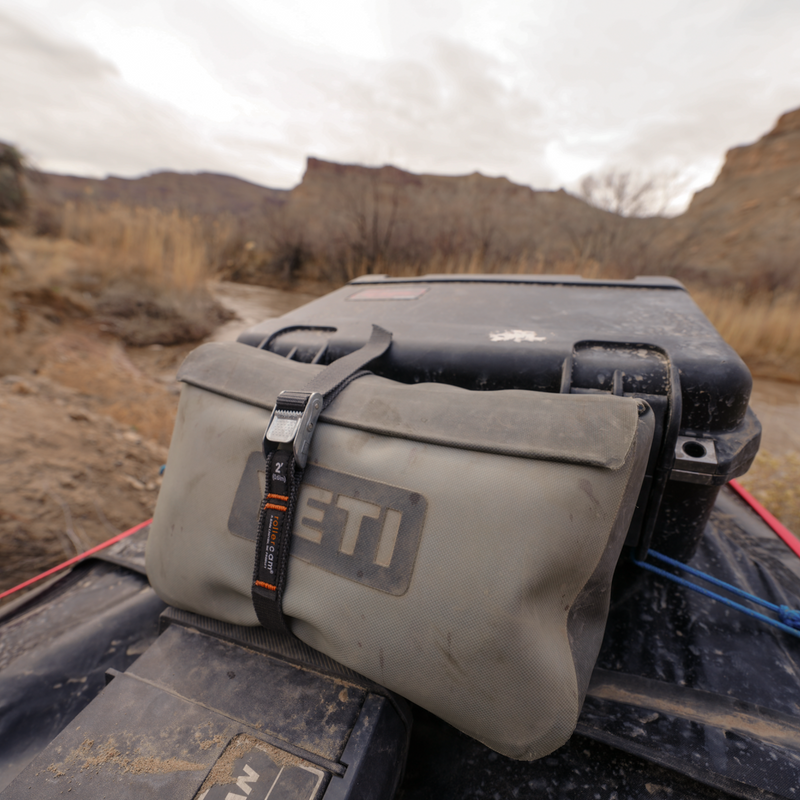 A gray YETI wet bag pouch strapped to a black hard case using a Rollercam 2ft straight end strap, outdoors in a desert canyon setting under an overcast sky.